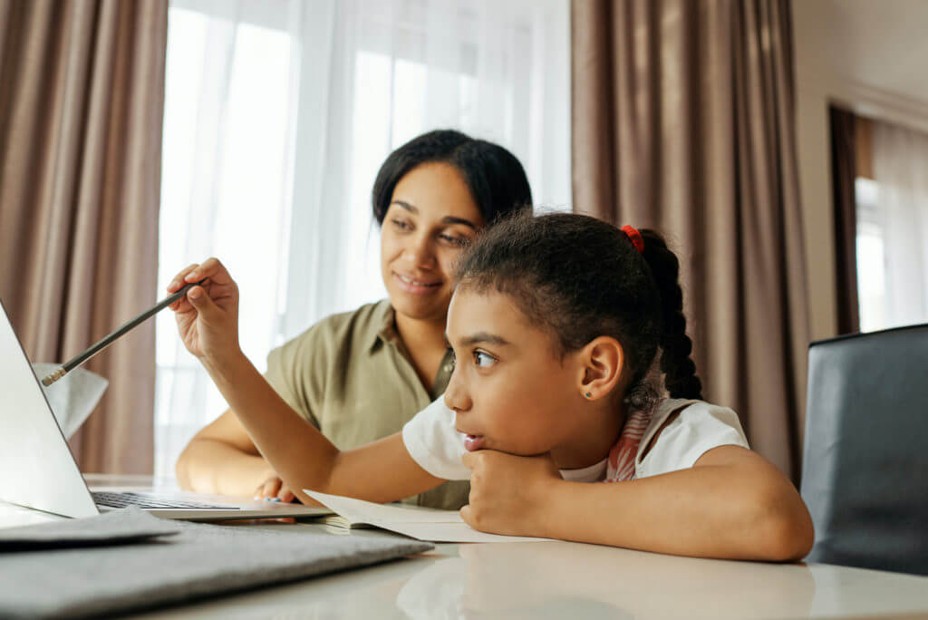 Young girl participating in remote learning with her mother looking on.