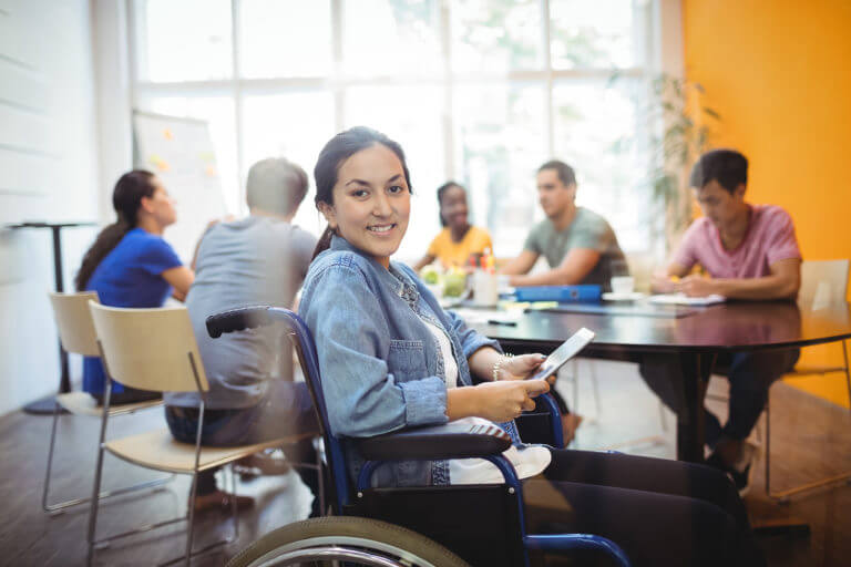 Young female adult, smiling, seated in a wheelchair and using digital tablet. Behind is a group of professionals working at a conference table.