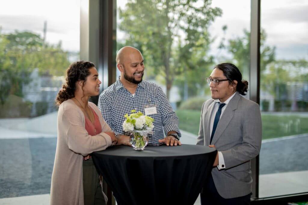 A professional chats with two students who are dressed professionally at an in-person networking event.